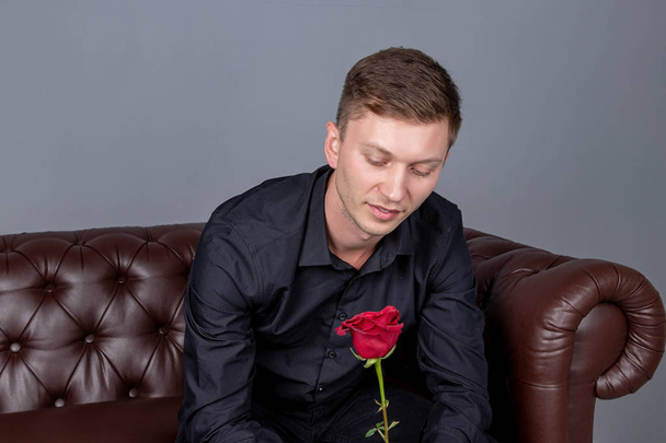 Handsome man wearing black shirt sitting on the leather sofa holding a rose and getting ready romantic date - Photo, Image