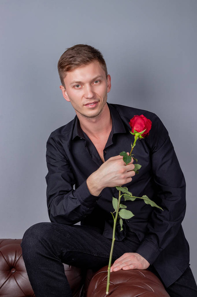 Handsome man wearing black shirt sitting on the leather sofa holding a rose and getting ready romantic date - Foto, afbeelding