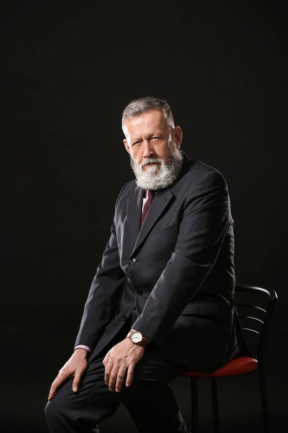 Handsome mature businessman sitting on chair against dark background - Foto, afbeelding