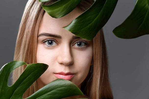 Όμορφη νεαρή γυναίκα με πλαστικοποιημένο βλεφαρίδες και τροπικά φύλλα σε γκρίζο φόντο - Φωτογραφία, εικόνα
