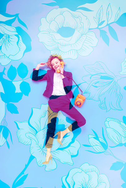 vista superior de la mujer de negocios en traje y zapatos de tacón alto hablando por teléfono mientras está acostado sobre fondo azul con ilustración floral
 - Foto, Imagen
