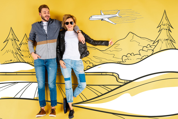 glückliche Frau zeigt Daumen hoch und umarmt Freund mit Bergen und Flugzeug-Illustration auf gelbem Hintergrund - Foto, Bild