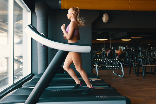  Blonde jonge, gemotiveerde vrouw die op de loopband in de sportschool loopt  - Foto, afbeelding
