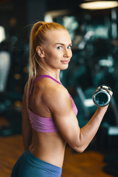 Young woman in pink top and mini shorts doing exercise with dumbbells while training arms at the gym  - Fotografie, Obrázek