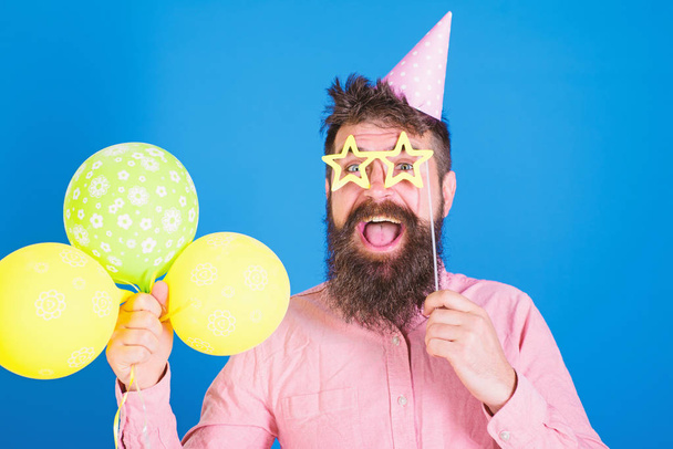 Happy bearded man in birthday cap with colourful baloons and paper star shaped glassses. Comedian with long beard and open mouth performing show. Bearded man entertaining guests at party, fun concept - Φωτογραφία, εικόνα