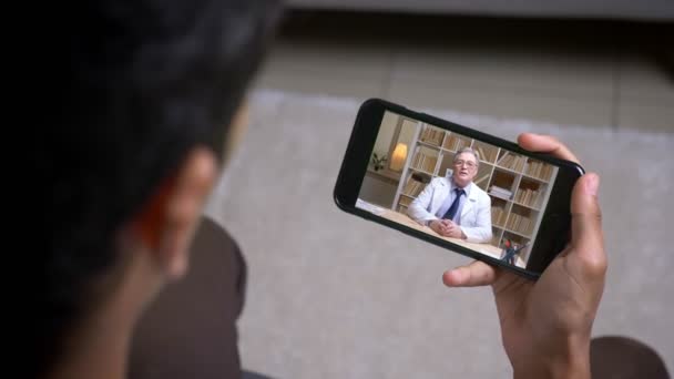 On-line videokall přes smartphone staršího lékaře v bílém laboratorním kabátě a s pacientem, který mluví vážně, za obrazovkou. - Záběry, video
