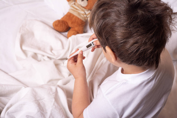 Больной ребенок с лихорадкой и болезнью в постели проверяет температуру термометром
. - Фото, изображение