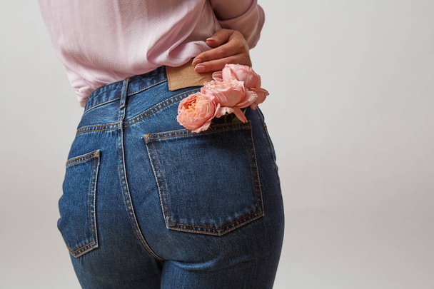 Perfect woman's bottom in a blue jeans and fresh roses living coral color in a back pocket on a light gray background, place for text. Concept of Woman's or Mother's Day. - Photo, image