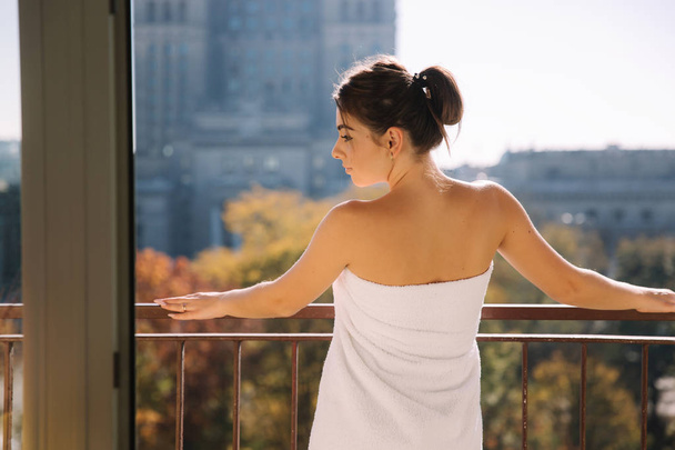Retrato de mujer sexy en toalla de baño de pie en el balcón. Los rayos del sol caen sobre mujer elegante
 - Foto, Imagen