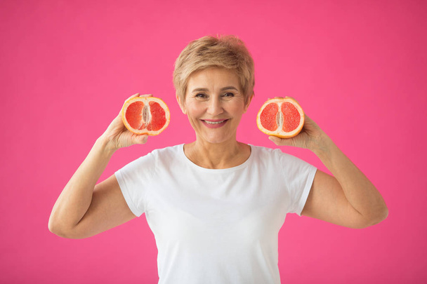 beautiful elderly woman in white T-shirt with grapefruits in her hands on a pink background - Valokuva, kuva