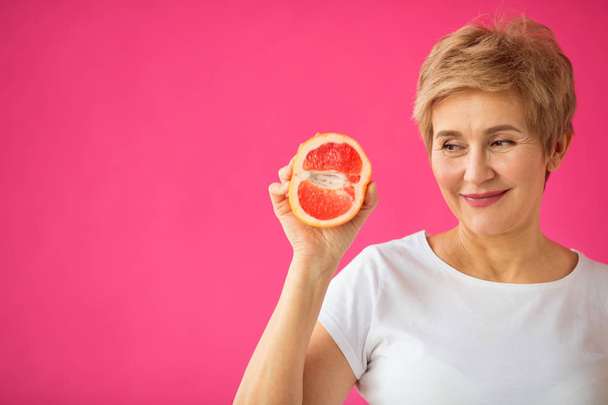 mooie oudere vrouw in wit T-shirt met grapefruits in haar handen op een roze achtergrond - Foto, afbeelding