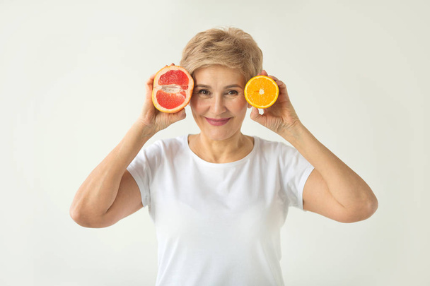 beautiful elderly woman in white t-shirt with orange and grape-fruit in hands on white background - Foto, afbeelding
