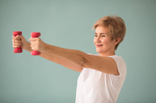beautiful elderly woman in white T-shirt goes in for sports with dumbbells on a blue background - Foto, Imagen