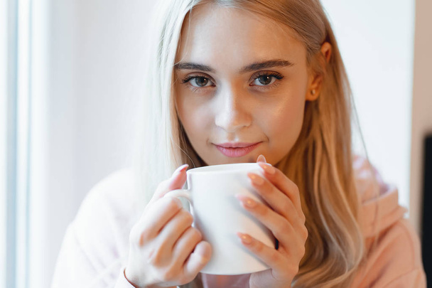 Skřítek, mladá dívka požívající šálek kávy nebo mléka, když sedí u okna uvnitř - Fotografie, Obrázek