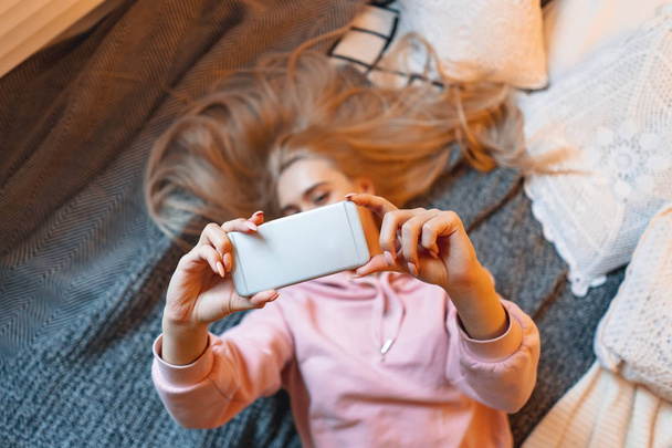 Όμορφη νεαρή κοπέλα που διασκεδάζει, κάνοντας selfie με έξυπνο τηλέφωνο, ενώ ξαπλωμένοι στο κρεβάτι στο σπίτι - Φωτογραφία, εικόνα
