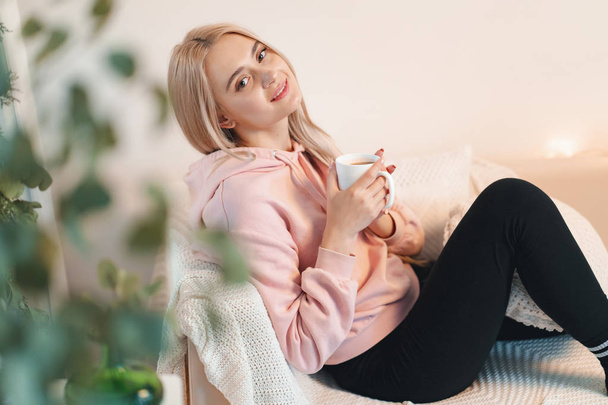Thoughtful young woman sitting on sofa with a cup of milk, coffee while relaxing at home - Фото, изображение