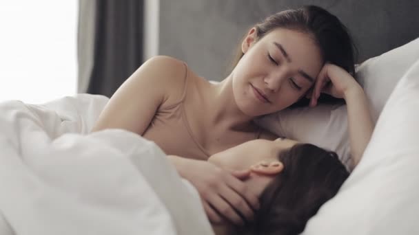 Lesbian couple hugging and smiling while lying together in bed at home. Young lesbians kisses and hugs after wake up - Кадры, видео
