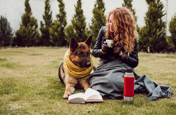Happy young woman holding thermos cup and sitting with her German shepherd dog on the grass, outdoor - Photo, image