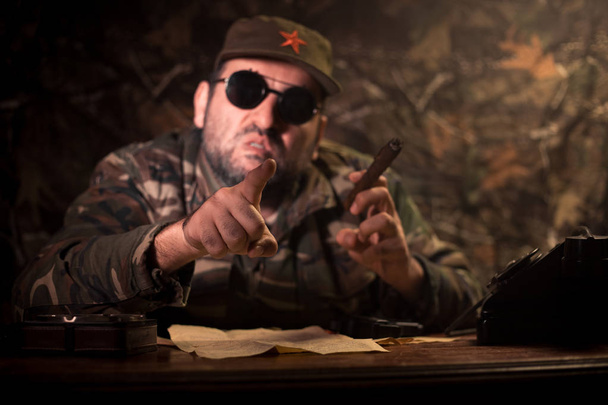 The evil dictator sitting on table. Angry communist general sitting at headquarter or Cuban commander in dark room. Studio decoration - Photo, Image
