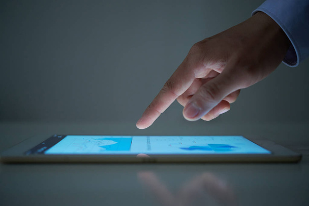 Рука анонимного бизнесмена прокручивает диаграммы на современном планшете в темной комнате
 - Фото, изображение
