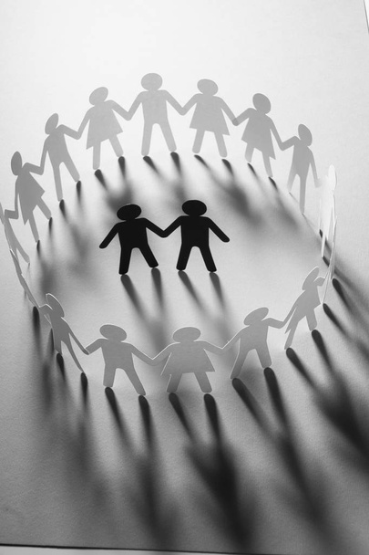 Figura de papel de pareja masculina rodeada por un círculo de personas de papel cogidas de la mano en la superficie blanca. Bulling, minorías, concepto de conflicto
. - Foto, imagen