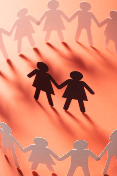 Paper figure of female couple surrounded by circle of paper people holding hands on red surface. Bulling, minorities, conflict concept. - Photo, Image