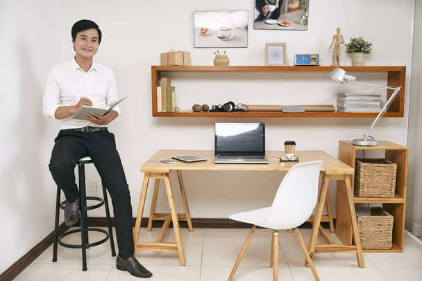 Retrato de sorrir jovem empresário asiático sentado nas fezes e escrever agenda em seu planejador
  - Foto, Imagem