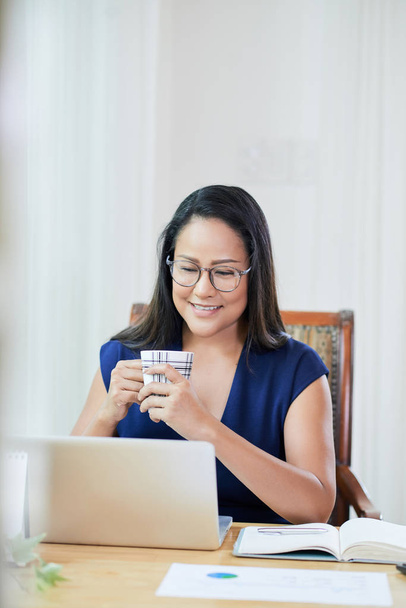 fröhliche asiatische Geschäftsfrau trinkt Morgenkaffee und checkt E-Mails auf ihrem Laptop - Foto, Bild