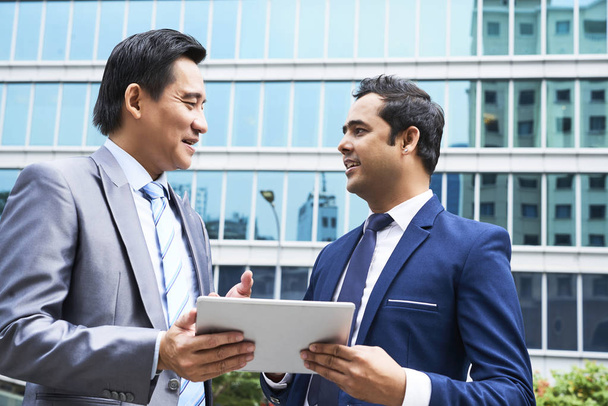Dois colegas de negócios em pé ao ar livre e discutindo apresentação em tablet digital com prédio de escritórios moderno em segundo plano
  - Foto, Imagem