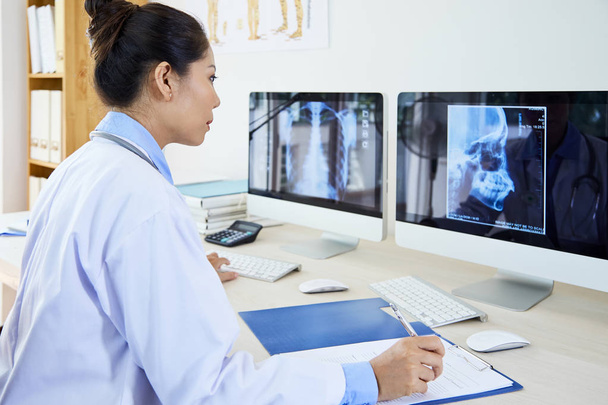 Médico asiático serio con bata de laboratorio sentado en su lugar de trabajo y examinando los escáneres de rayos X en los monitores de computadoras y defectos de escritura en el documento
 - Foto, imagen