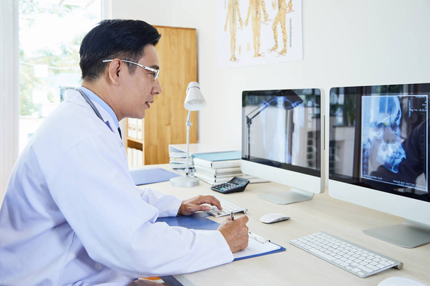 Asiatique mature neurologue dans les lunettes examiner l'image de rayons X sur écran d'ordinateur et de prendre des notes sur presse-papiers tout en travaillant à son lieu de travail
 - Photo, image