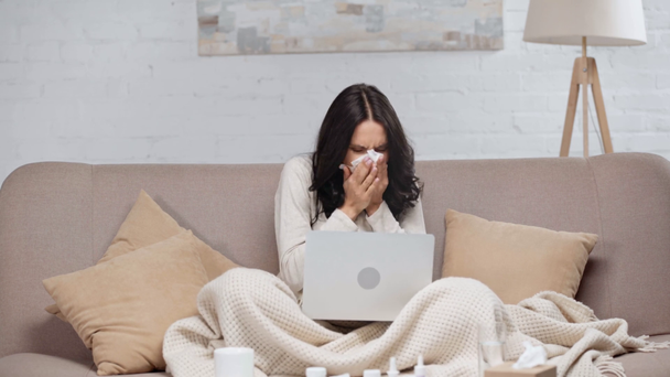 brünette kranke Bloggerin benutzt Laptop, hält Tasse, trinkt Tee, nimmt Gewebe und niest im Wohnzimmer  - Filmmaterial, Video
