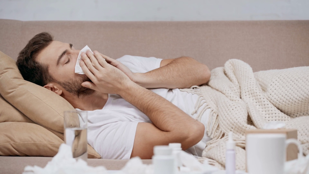 άρρωστος και όμορφος άντρας ξαπλωμένος κάτω από κουβέρτα, παίρνοντας ιστό και φυσώντας τη μύτη στο σαλόνι  - Πλάνα, βίντεο