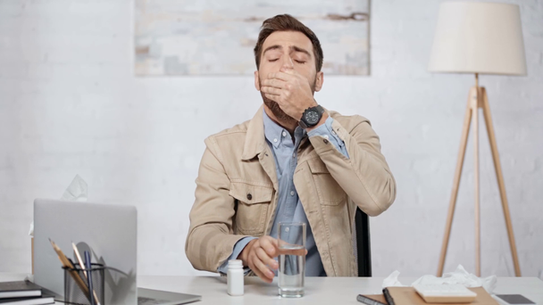 pohledný vousatý muž kýchání, pohled na láhev, prášky a pitná voda poblíž laptopu v kanceláři  - Záběry, video