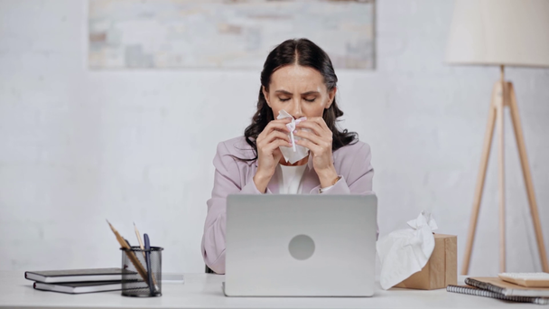 Ελκυστική μελαχρινή επιχειρηματίας χρησιμοποιώντας φορητό υπολογιστή, φτάρνισμα, παίρνοντας πετσέτα από το κουτί ιστού ενώ κάθονται στο γραφείο  - Πλάνα, βίντεο