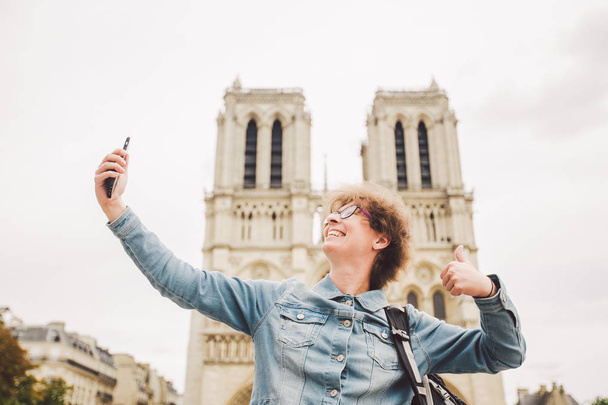 Tourist in Paris making funny selfie near Notre Dame Cathedral. Beautiful young Caucasian tourist woman with backpack in Paris making funny selfie hand holding phone, photo near Notre Dame Cathedral - Photo, image