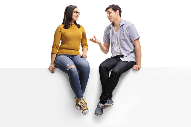 Full length shot of a young man sitting on a blank signboard and talking to a young woman isolated on white background - Foto, immagini