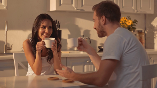 cheerful brunette woman holding cup, talking with man, gesturing, drinking coffee and laughing in kitchen  - Záběry, video