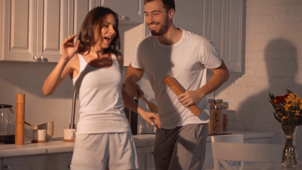 cheerful brunette woman dancing with happy bearded man holding wooden spoon and pepper shaker in kitchen  - Felvétel, videó