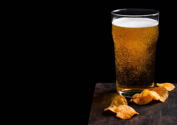 Glass of lager beer with potato crisps snack on vintage wooden board on black background. Beer and snack. Space for text - Foto, Bild