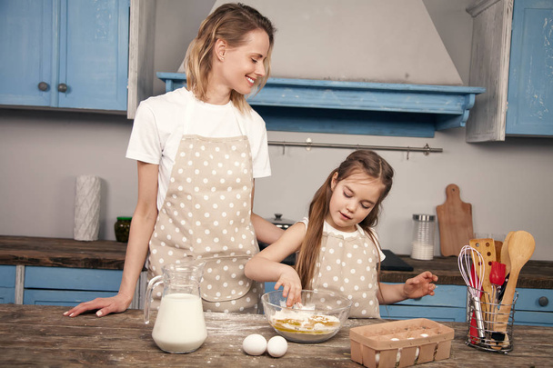 Η μαμά και η μικρή κόρη στην κουζίνα ετοιμάζουν ζύμη, Ψήνοντας μπισκότα. Η μαμά διδάσκει στην κόρη της να ζυμώνουν τη ζύμη από αλεύρι, γάλα και αυγά. Μικρή βοηθός. Σπιτικό φαγητό - Φωτογραφία, εικόνα