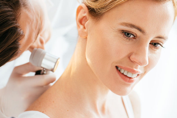 selective focus of attractive woman smiling while dermatologist examining neck with dermatoscope  - Foto, imagen