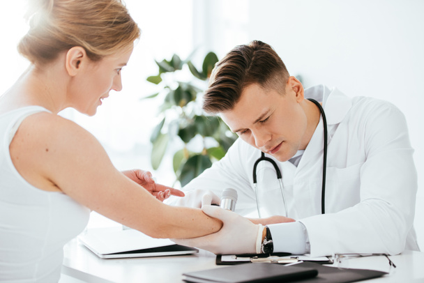 guapo dermatólogo examinando mano de atractiva mujer mientras sostiene dermatoscopio
  - Foto, Imagen