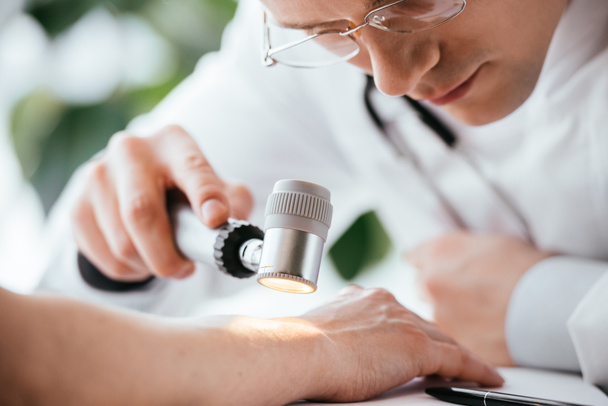 επιλεκτική εστίαση του δερματολόγου σε γυαλιά που κατέχουν δερματοσκόπιο κοντά στο χέρι του ασθενούς  - Φωτογραφία, εικόνα
