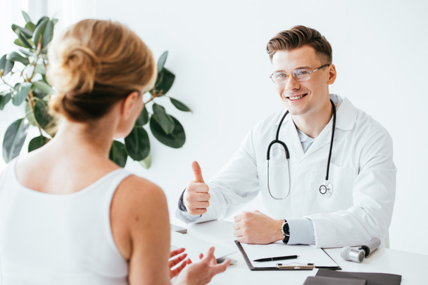 selective focus of cheerful doctor in glasses showing thumb up while looking at patient  - Фото, зображення