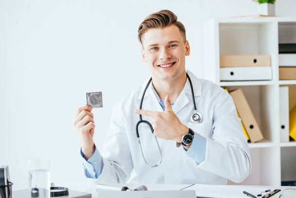 όμορφος και χαρούμενος γιατρός σε λευκό παλτό δείχνοντας με το δάχτυλο στο προφυλακτικό - Φωτογραφία, εικόνα