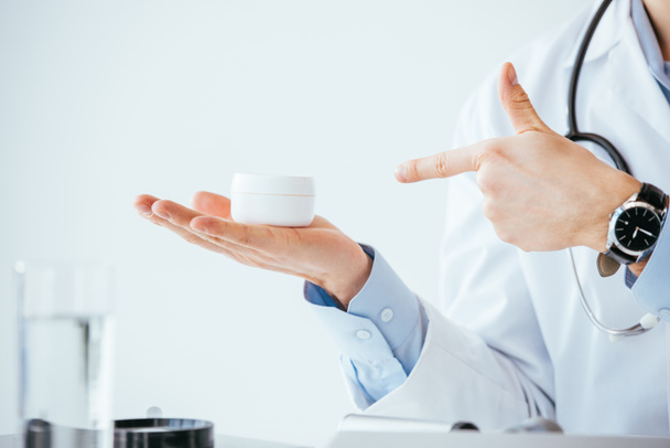 cropped view of doctor in white coat pointing with finger at container with cosmetic cream  - Fotoğraf, Görsel