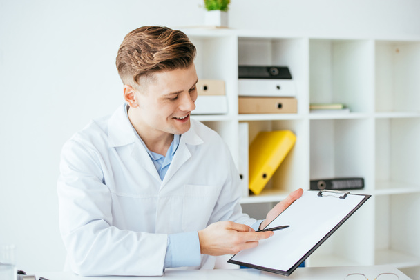 fröhlicher Arzt in weißem Mantel mit Stift und Blick auf leeres Klemmbrett  - Foto, Bild
