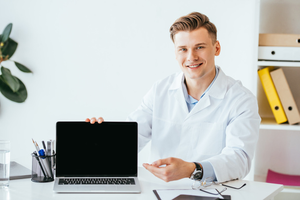 cheerful doctor in white coat sitting near laptop with blank screen  - Foto, afbeelding