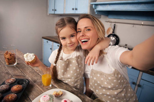szczęśliwy kochający rodzina w kuchni. Matka i dziecko córka dziewczyny jedzą ciasteczka, które zrobiły i bawią się w kuchni. Domowe jedzenie, mały pomocnik. Matka robi selfie siebie i - Zdjęcie, obraz
